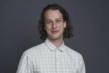 Professional headshot of Benjamin Kasavan. He is wearing a button up shirt. There is a gray background.